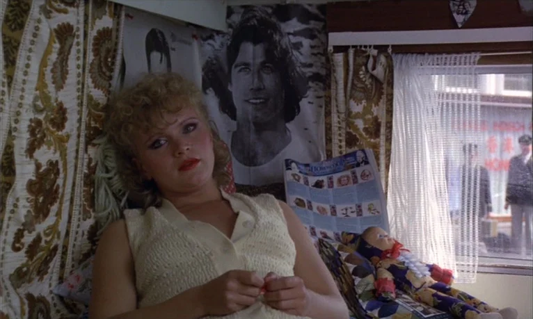 Spetters (1980) movie still: medium — A cramped caravan interior decorated with floral curtains and a John Travolta poster. A young blonde woman…