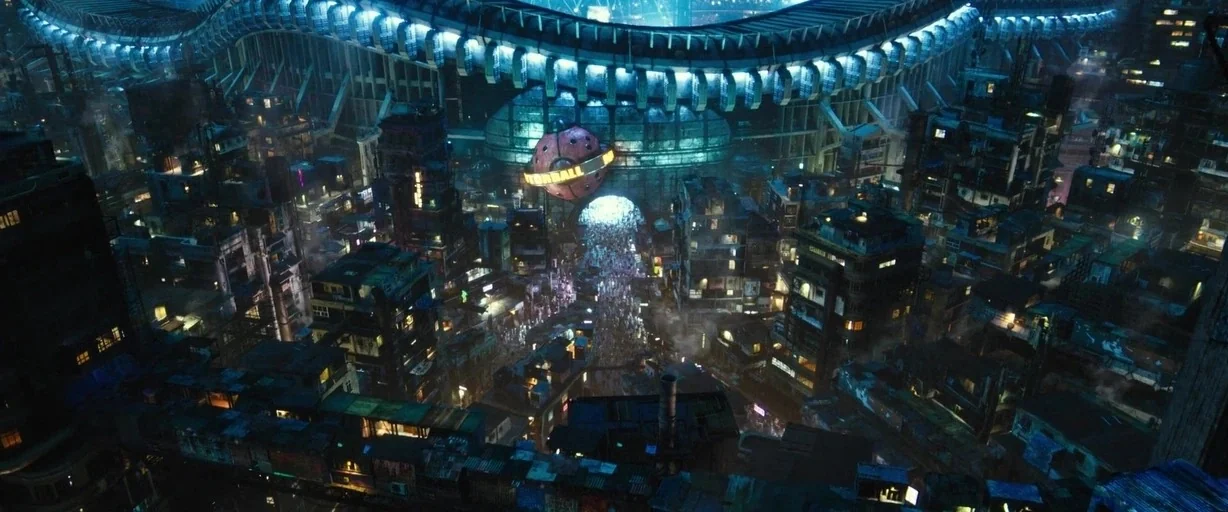 Alita: Battle Angel movie still: establishing — A dense, futuristic slum spans a narrow valley between sprawling, dark, multi-story apartment…