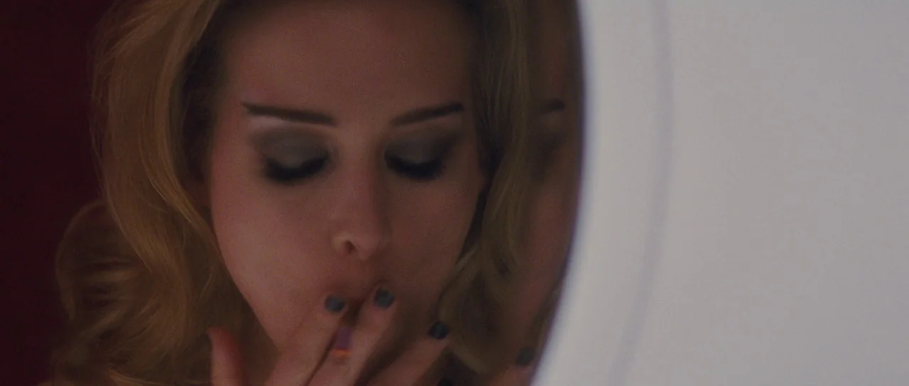 Looper (2012) movie still: closeup — A close-up view of a woman looking at her reflection in a circular mirror. She holds a lit cigarette to her…
