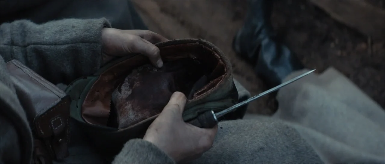 The Rifleman (2019) movie still: closeup — A mud-filled trench environment with dark, damp earth visible on the ground. A pair of hands in woolen…