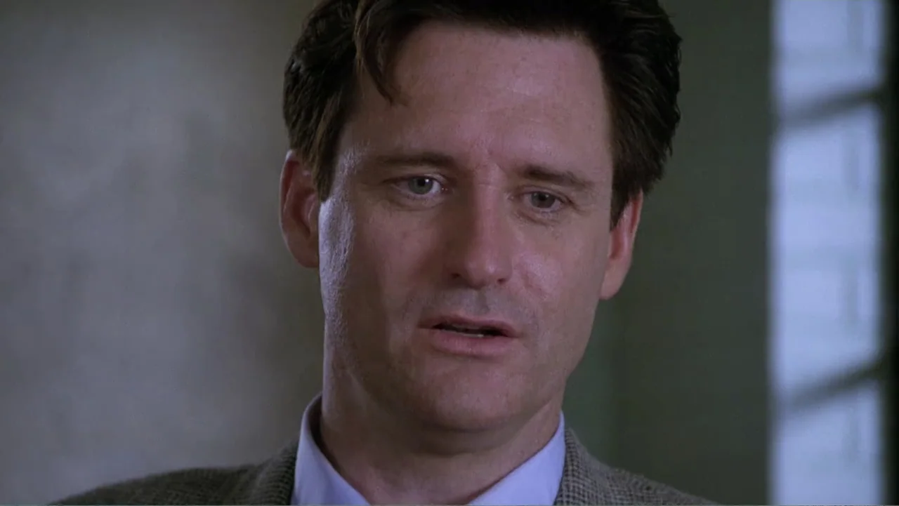 Mistrial (1996) movie still: closeup — A dimly lit indoor office space with blurred structural elements in the background. A man in a collared shirt…