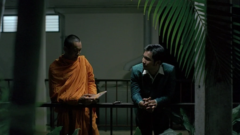 Syndromes and a Century (2006) movie still: medium — A dimly lit concrete balcony at night with palm leaves framing the foreground. Two men stand by…