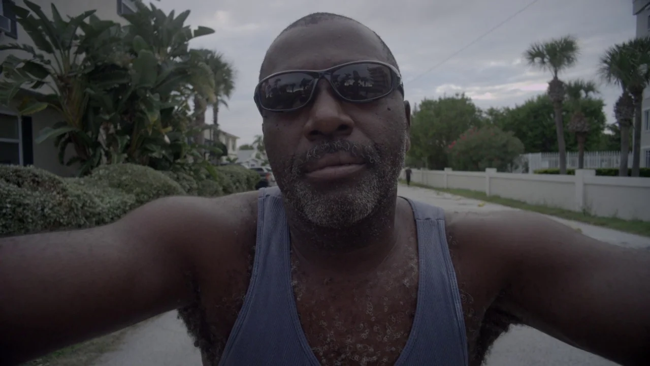 Florida Man (2015) movie still: closeup — An outdoor path lined with tropical vegetation and white walls under an overcast sky. A man in a tank top…