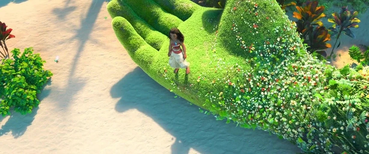 Moana (2016) movie still: wide — A giant green hand covered in small flowers rests on a sandy beach. A young girl stands on the palm of the giant…