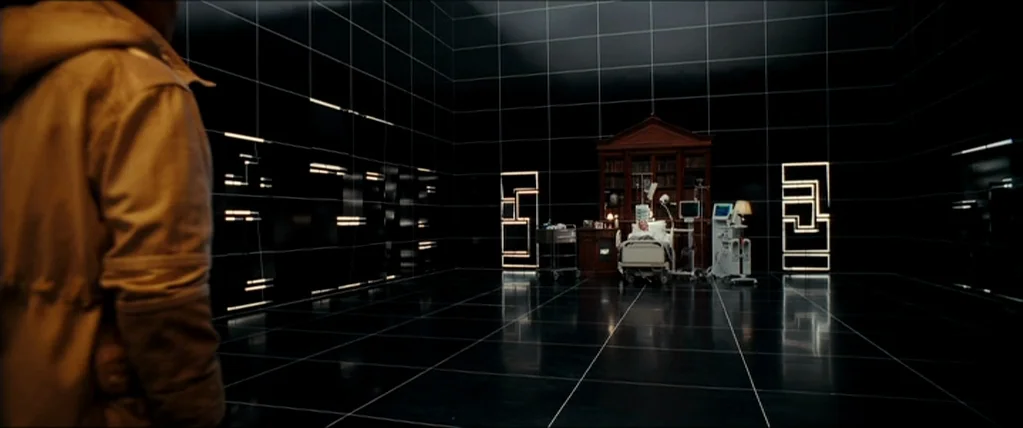 Inception (2010) movie still: wide — A vast black room featuring glowing white grid lines on all surfaces. A hospital bed and wooden desk are…