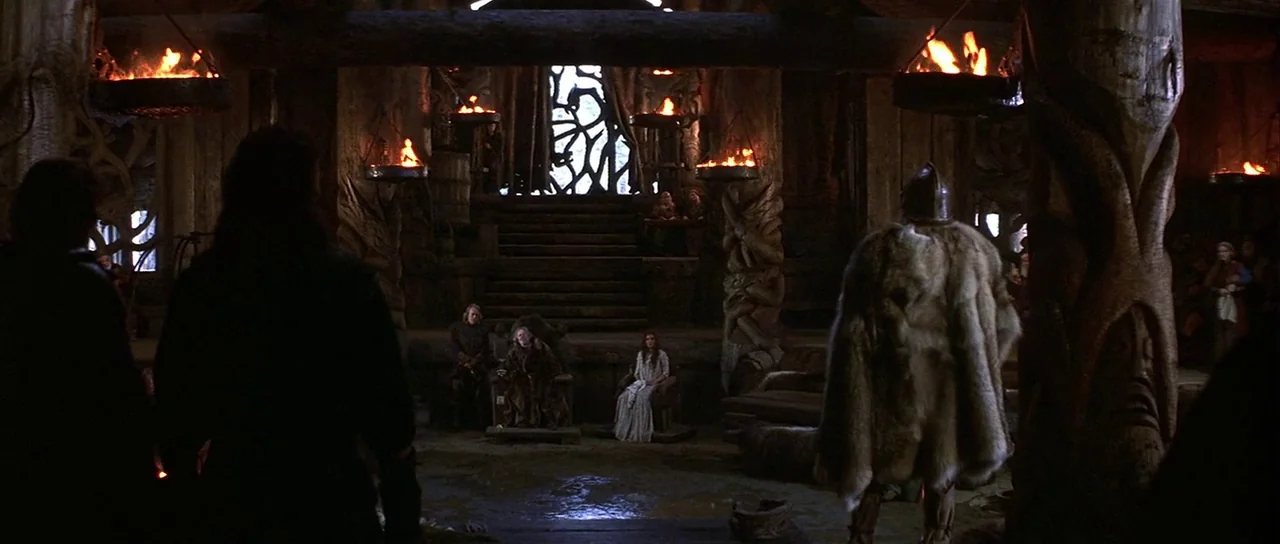 The 13th Warrior (1999) movie still: wide — A rustic wooden hall features carved pillars and elevated seating for a ruling figure. A cloaked man…