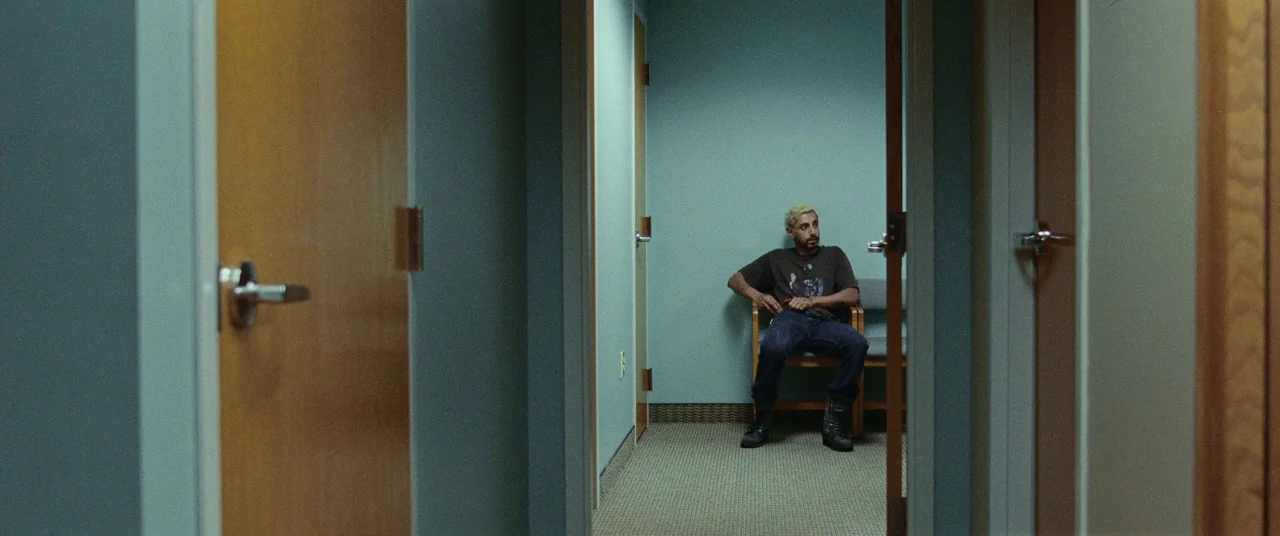 Sound of Metal (2019) movie still: wide — A sterile, narrow institutional hallway with light blue walls and wood-trimmed doorways. A man sits alone…