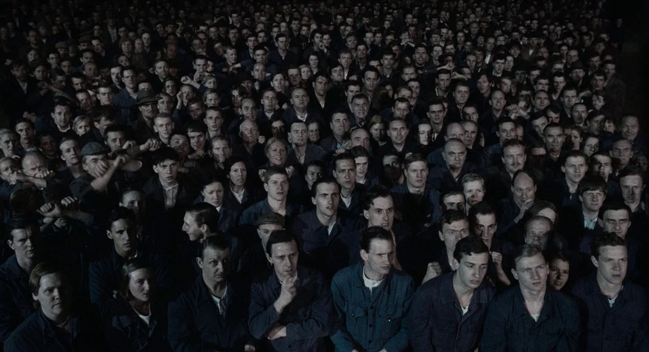 1984 (1984) movie still: wide — A dense crowd of men in work uniforms stand together in a large room. The people are packed tightly and look toward…