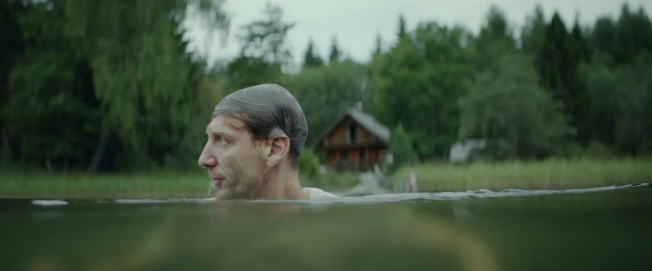 Dogs Don't Wear Pants (2019) movie still: medium — A calm lake surrounded by dense green forest under an overcast sky. A man swims while a small…