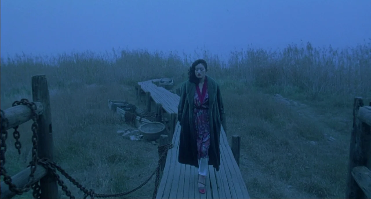Shanghai Triad (1995) movie still: fullbody — A wooden pier extends into a misty, grassy wetland area during the blue hour. A woman walks slowly…