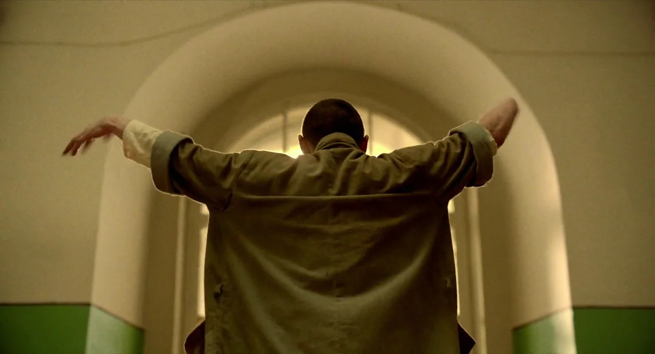Beanpole (2019) movie still: medium — An interior hallway features a large arched window that fills the frame with light. A man with a shaved head…