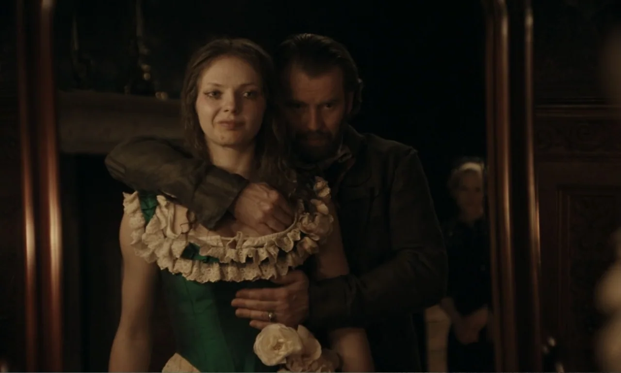 The Ugly Stepsister (2025) movie still: medium — An interior space with period furniture and a large mirror reflecting the two subjects. A man…