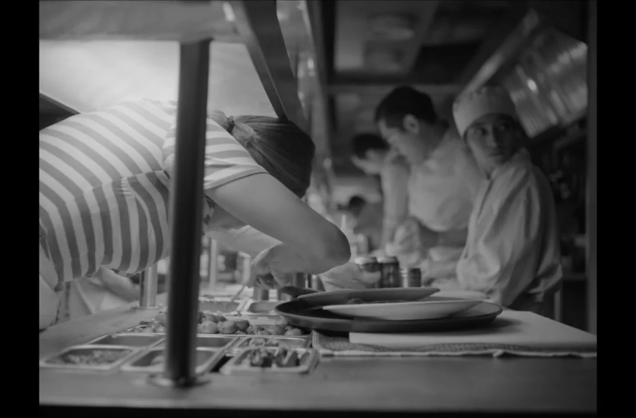 The Kitchen (2012) movie still: medium — A busy professional kitchen station with stainless steel counters and various food containers. A woman in a…