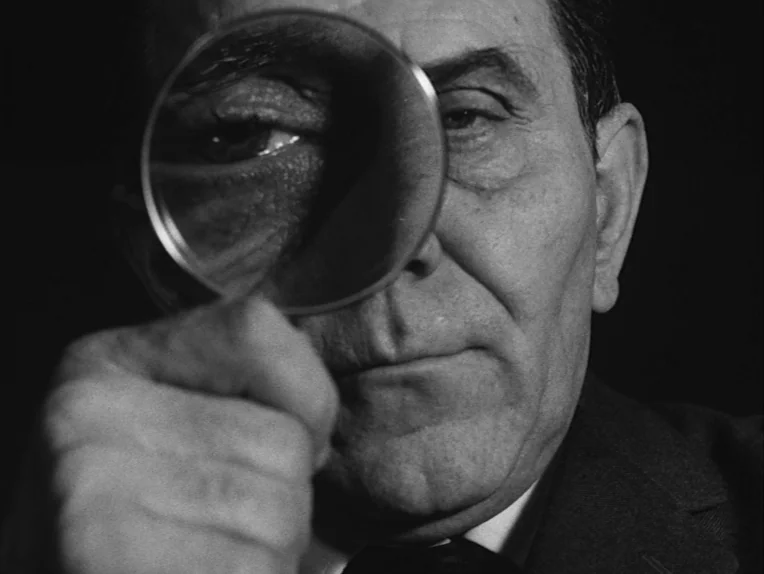 The Rite (1969) movie still: closeup — A man stands against a dark, featureless background. He holds a round magnifying glass directly in front of…