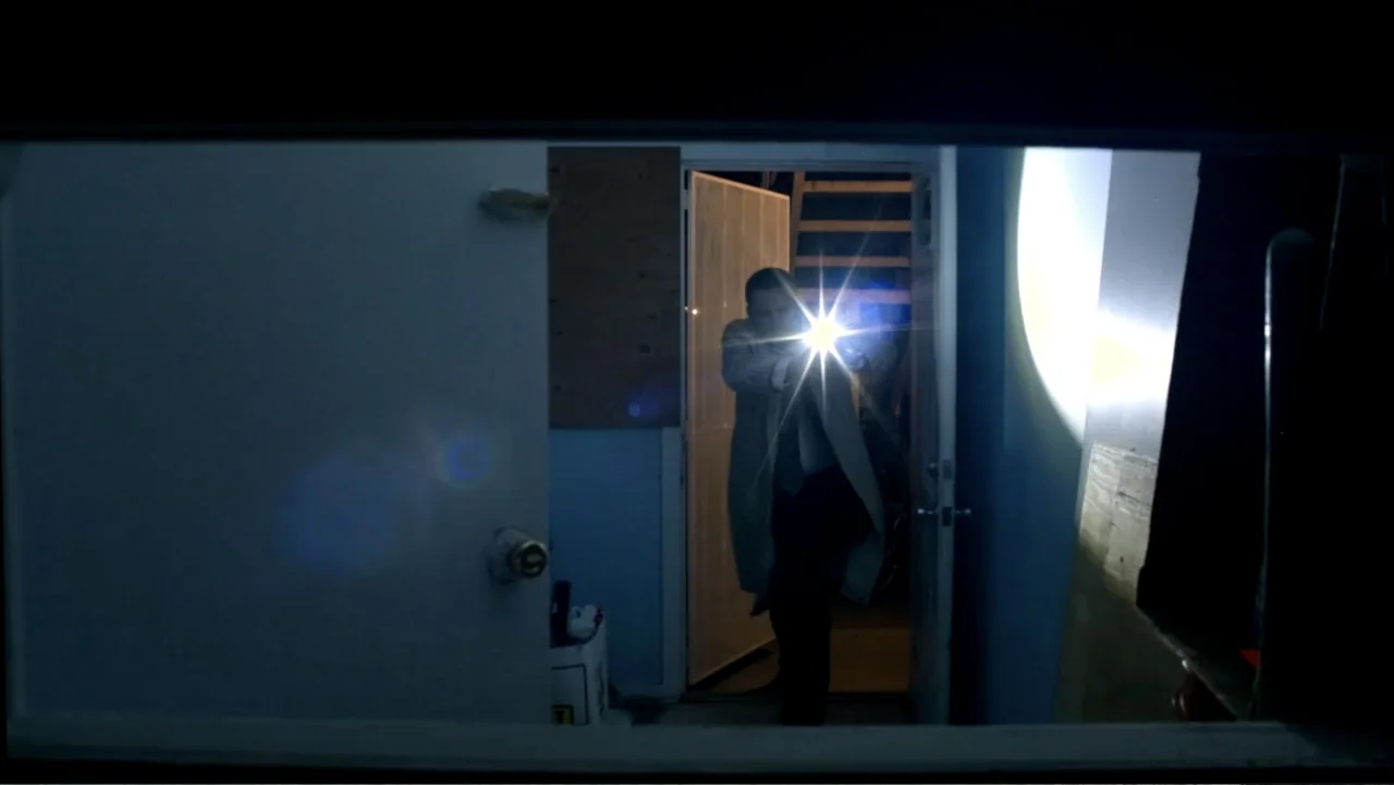 Abandoned Dead (2015) movie still: medium — A dark interior is viewed through an opening, revealing a man standing in a doorway. The man holds a…