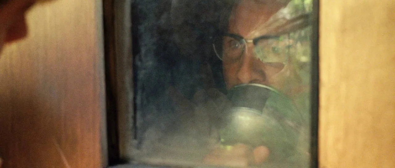 On The Road (2012) movie still: closeup — A man looks through a small square window set into a wooden frame partition. The man holds a circular lamp…