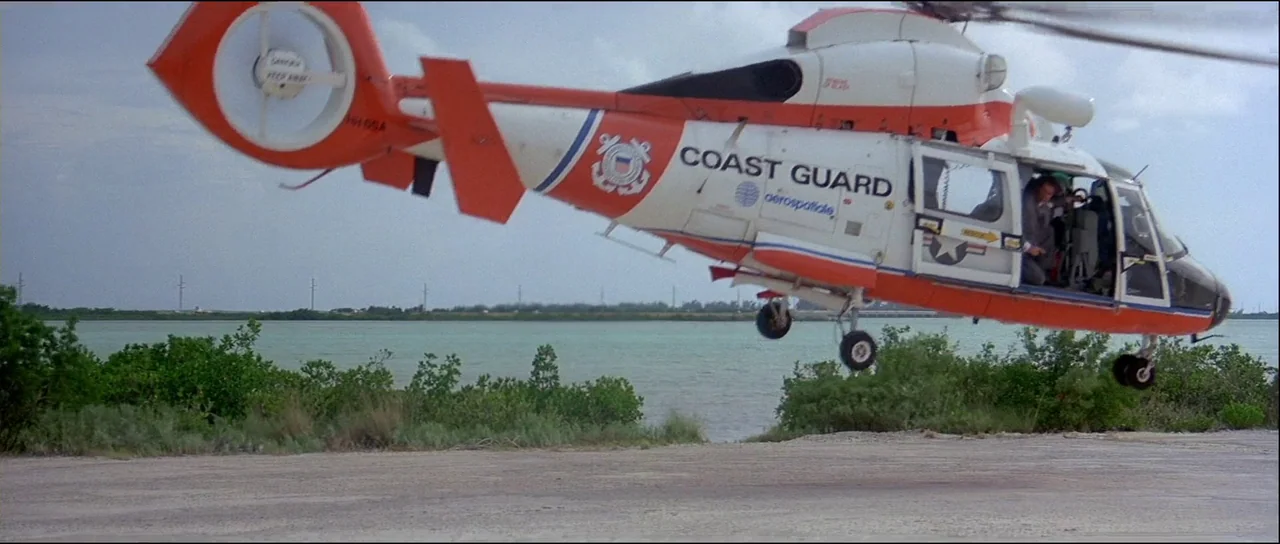 Licence to Kill (1989) movie still: wide — A United States Coast Guard helicopter hovers low over a gravel clearing near water. The pilot looks out…