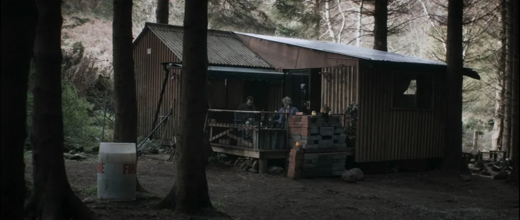 One Hundred Mornings (2009) movie still: wide — A wooden cabin sits surrounded by tall trees in a dense forest area. Two people sit on the deck of…