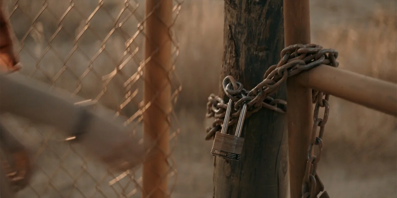Fall (2022) movie still: closeup — A metal chain and silver padlock wrap around a wooden post of a fence. The blurred silhouette of a person's hand…