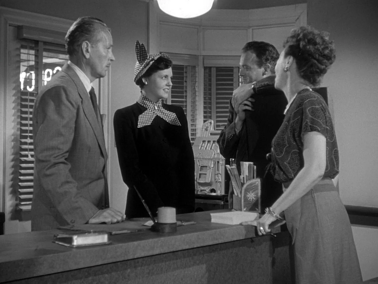 The Prowler (1951) movie still: medium — A hotel check-in counter is occupied by four people standing in a line. The group appears to be interacting…