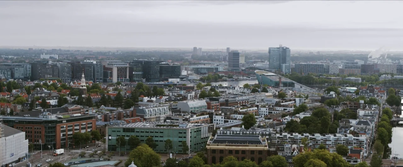 Black Lotus (2023) movie still: establishing — An aerial view of the Amsterdam skyline shows dense blocks of urban architecture. Numerous buildings…