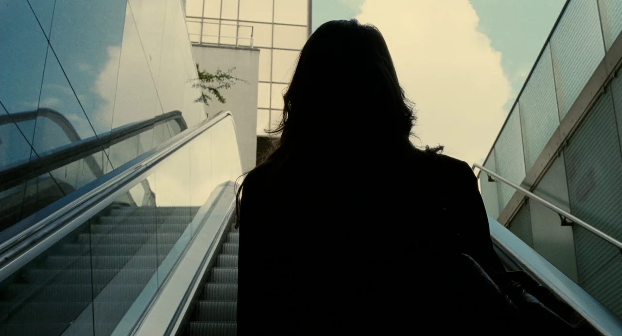 Young & Beautiful (2013) movie still: medium — An outdoor escalator rises toward the sky alongside a modern building with glass panels. A woman is…