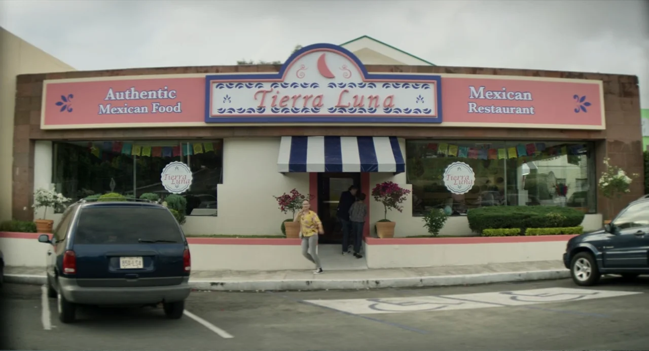 A Million Miles Away (2023) movie still: wide — The exterior of a Mexican restaurant called Tierra Luna with a pink and blue sign. A woman in a…