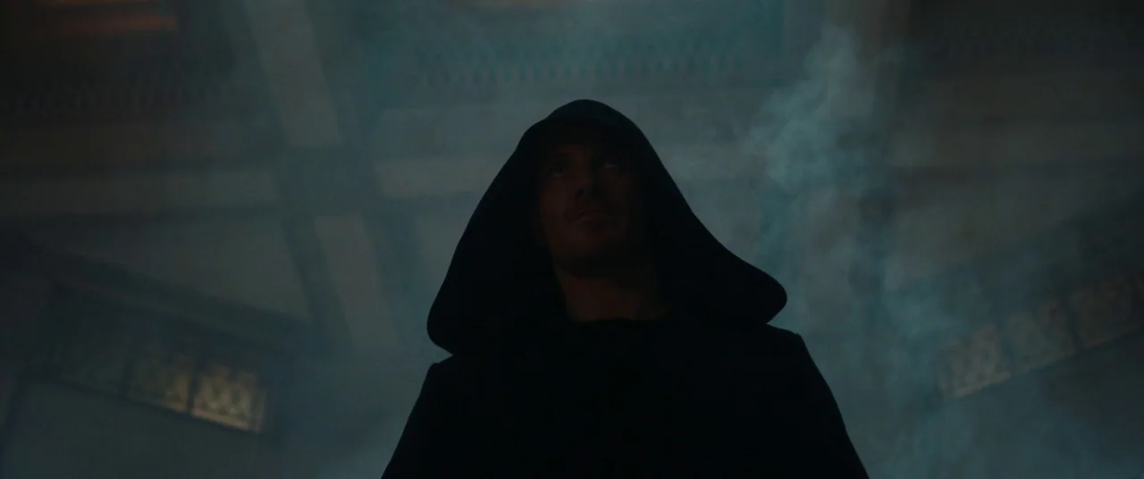 Assassin’s Creed (2016) movie still: medium — A dark hallway filled with thick atmospheric haze and low-level lighting. A man wearing a dark hood…