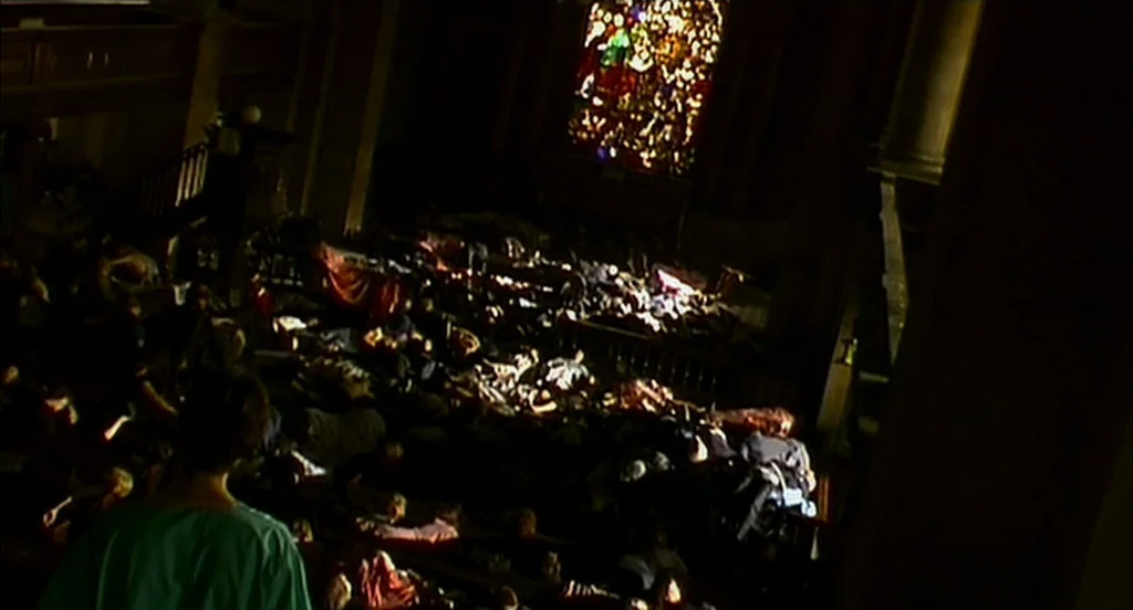 28 Days Later (2002) movie still: wide — A dark church interior is filled with a large, huddled crowd on the floor. A vibrant stained glass window…