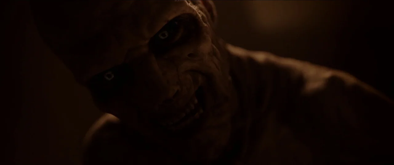 His House (2020) movie still: closeup — A dark, featureless environment frames an entity with decaying, textured skin. The subject gazes intensely…