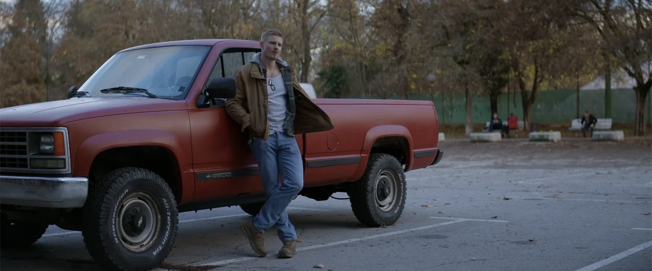 Marked Men: Rule + Shaw (2025) movie still: fullbody — A paved asphalt parking lot sits adjacent to a grove of autumn trees. A man in jeans and a…