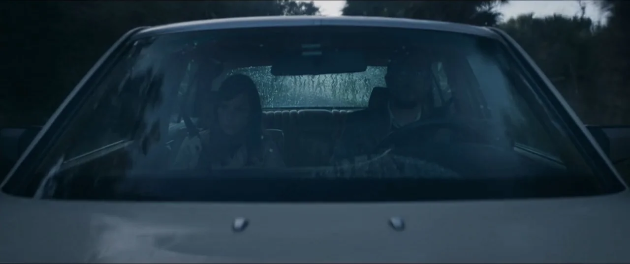 Offseason (2022) movie still: medium — A rain-covered windshield provides a view of two people sitting inside a moving car. A woman sits in the…