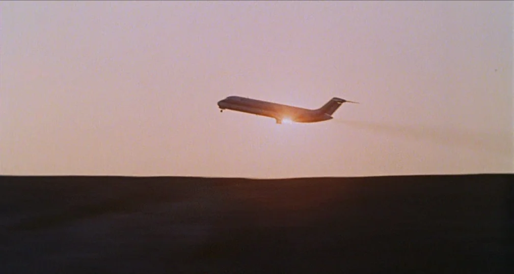 Midnight Cowboy (1969) movie still: wide — A silhouette of an airplane traverses a vast, empty expanse of sky during sunset. The low sun creates a…