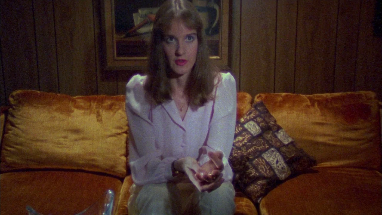 Poison movie still: medium — A room with brown wood-paneled walls features a mustard yellow velvet sofa. A young woman sits on the sofa looking…