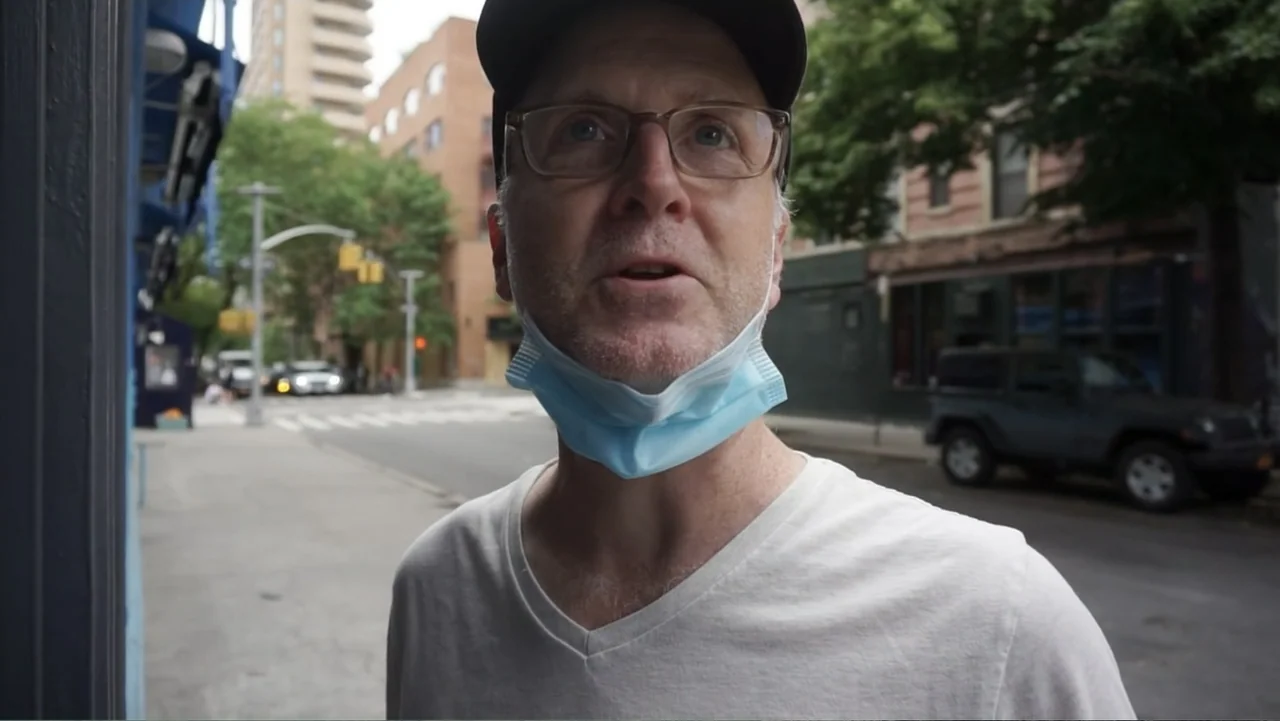 Covid Diaries NYC (2021) movie still: closeup — A suburban street corner with brick buildings and a parked black suv in background. A middle-aged…