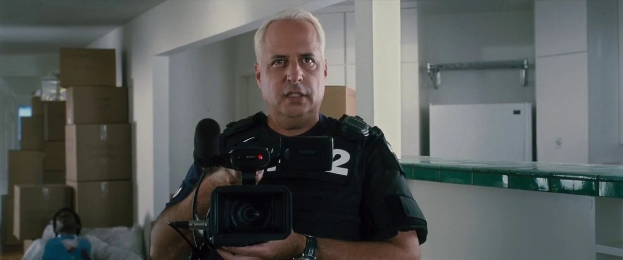 Southland Tales (2006) movie still: medium — A man stands inside a house filled with cardboard moving boxes. He holds a professional camcorder…