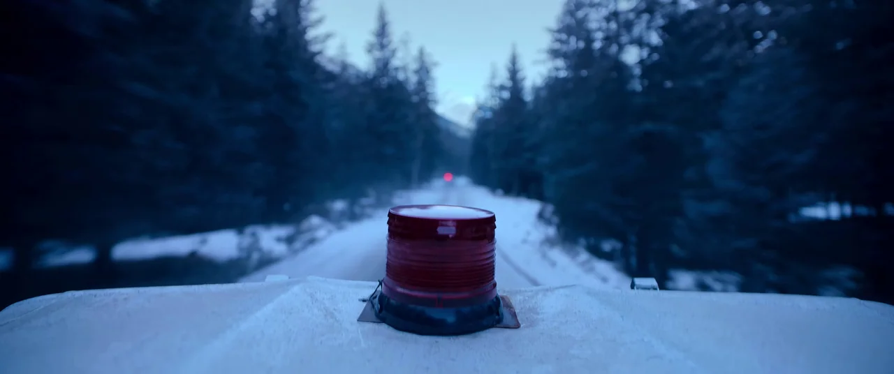 One And Four (2021) movie still: closeup — A red flashing beacon is mounted on a white surface overlooking a snowy road. Pine trees flank the icy…
