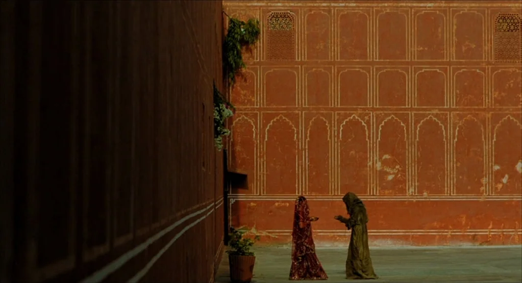 The Fall movie still: wide — A spacious courtyard features high, red walls with decorative arched moldings and white lines. Two women stand facing…