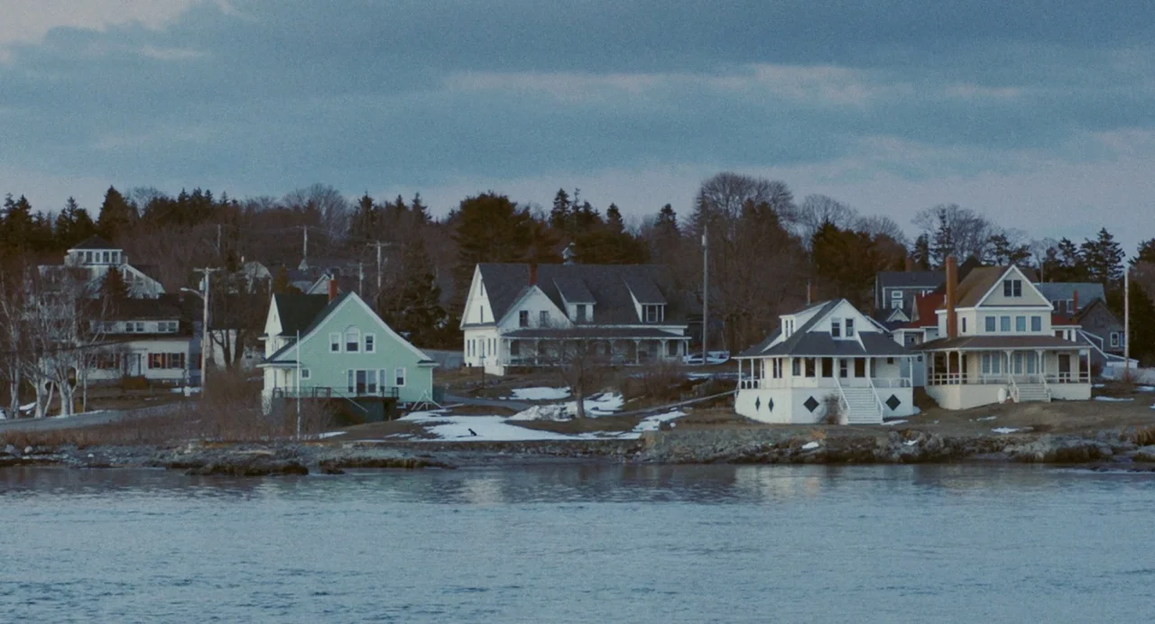 Blow The Man Down (2019) movie still: wide — A collection of wood-frame houses sits along a calm shoreline under an overcast sky. Patches of snow…