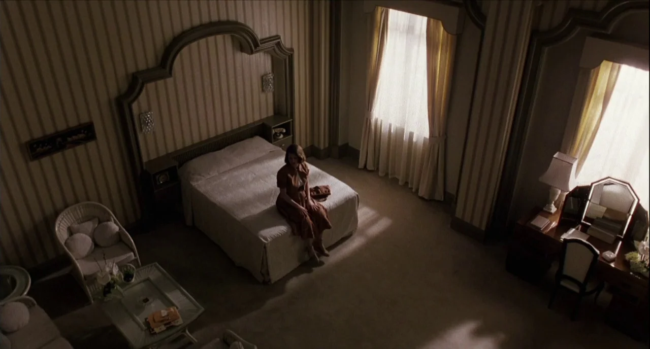 The Hours (2002) movie still: wide — A hotel room with vertical striped wallpaper, a large bed, and vanity furniture. A young woman sits alone on…