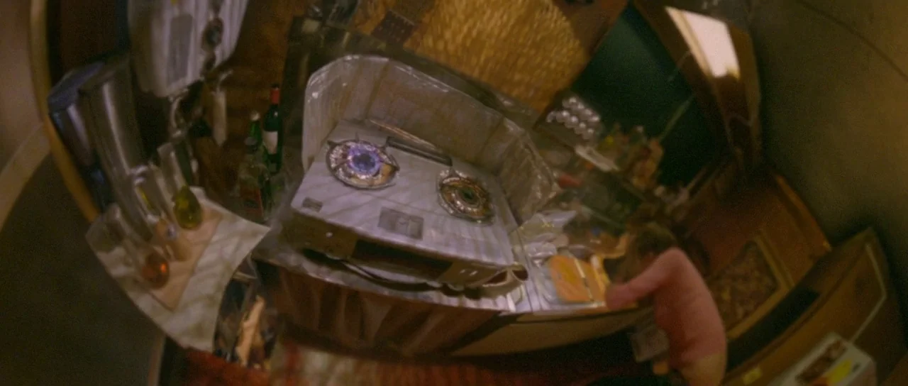 Enter The Void (2009) movie still: wide — A cramped kitchen with a portable gas stove ignited on the main counter. A woman in a pink shirt leans…