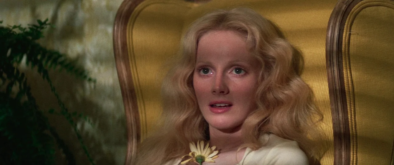 Death Game (1977) movie still: closeup — A woman sits in a yellow upholstered chair with a green fern visible nearby. She holds a small white daisy…