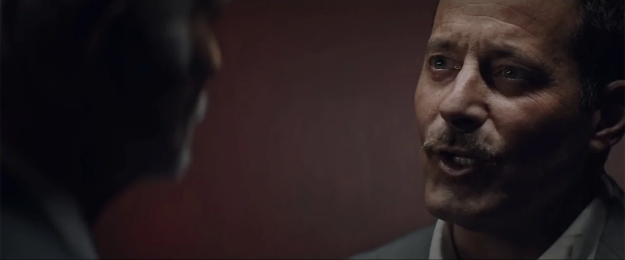 Gender Under Attack (2018) movie still: closeup — An interior office space with dim, dark-toned wall surfaces. A man in a suit speaks during a…