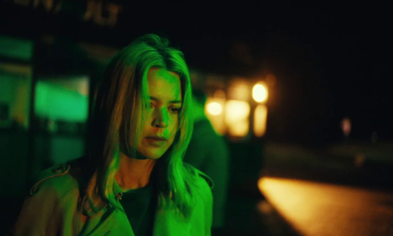 Just The Two Of Us (2023) movie still: medium — A dark outdoor space at night near a commercial building with green neon signage. A blonde woman in…