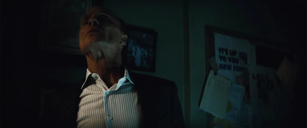 Run All Night (2015) movie still: medium — A dark office environment features a bulletin board with flyers on the wall behind. An older man in a…