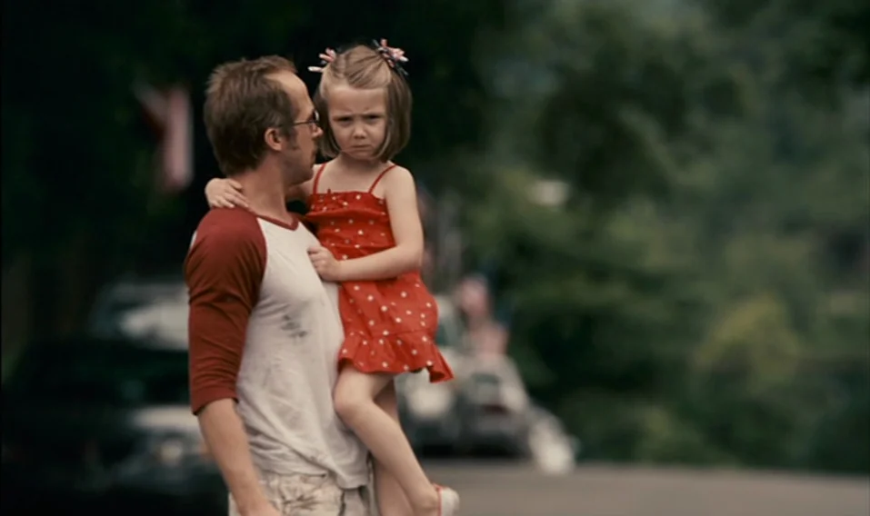 Blue Valentine (2010) movie still: medium — A man carries a small girl along a paved road lined with trees. The man holds the girl securely against…
