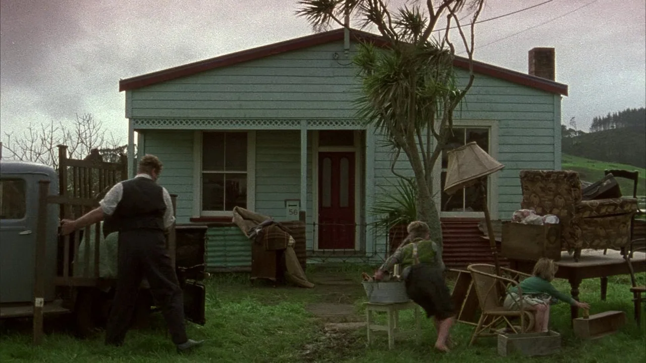An Angel At My Table (1990) movie still: wide — A pale green wooden house sits in a grassy yard under a cloudy sky. An adult man and two females are…