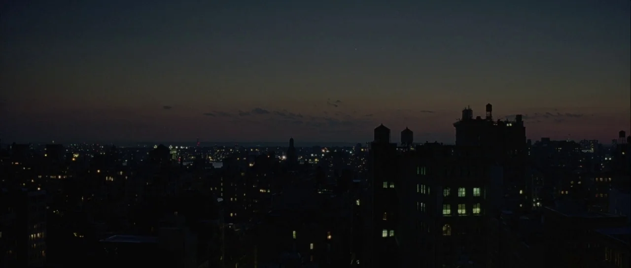 Capote (2005) movie still: establishing — A dark, expansive view of a city skyline at dusk with scattered interior lights. Silhouetted buildings…