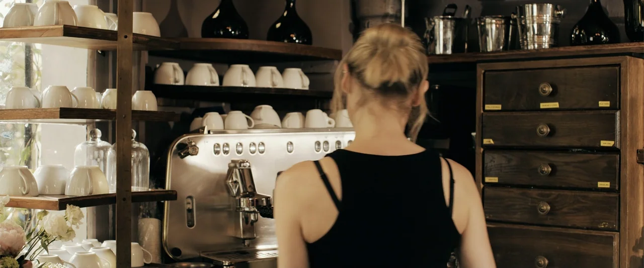 The 355 (2022) movie still: medium — A wooden shelving unit holds numerous white ceramic cups and dark glass bottles. A woman in a black tank top…