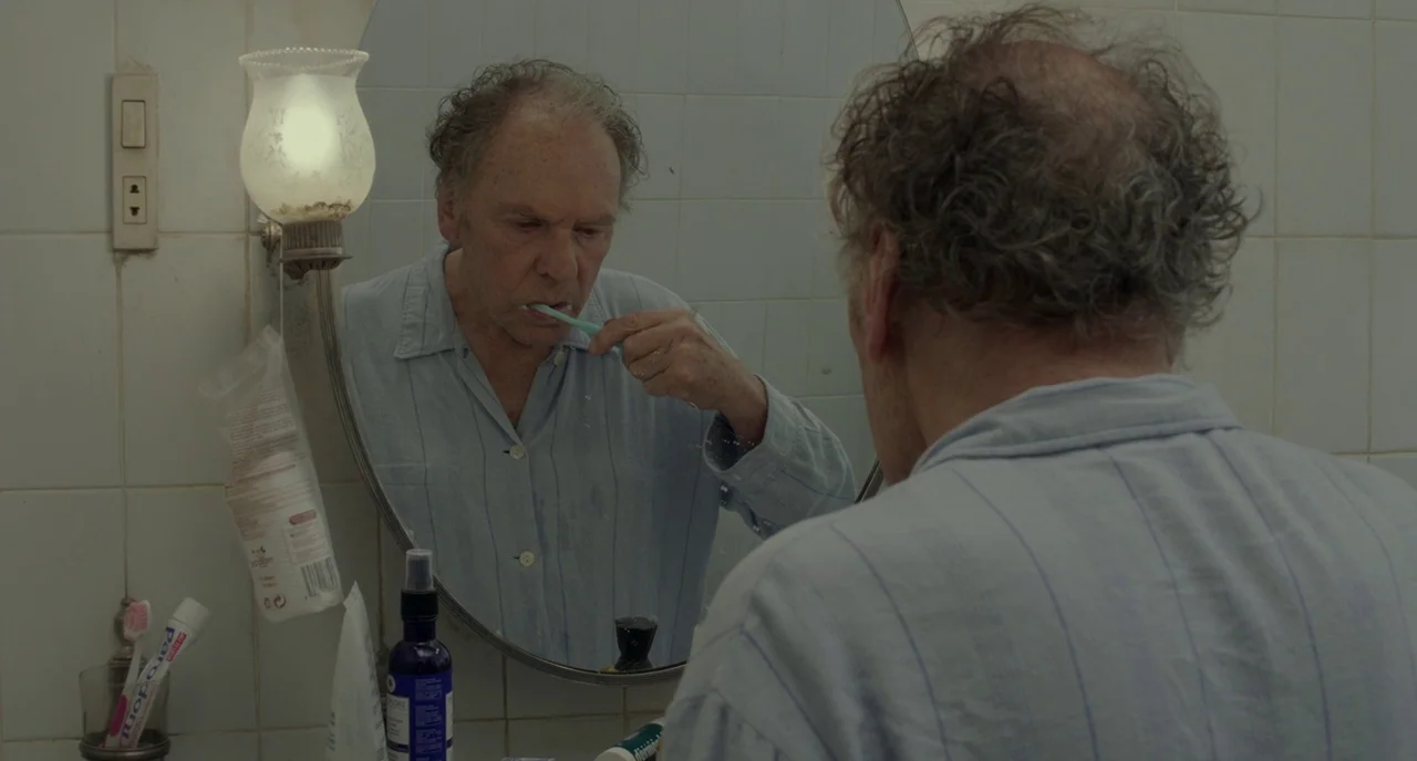 Amour (2012) movie still: medium — A tiled washroom with a wall-mounted light fixture and a circular mirror. An elderly man stands before the mirror…